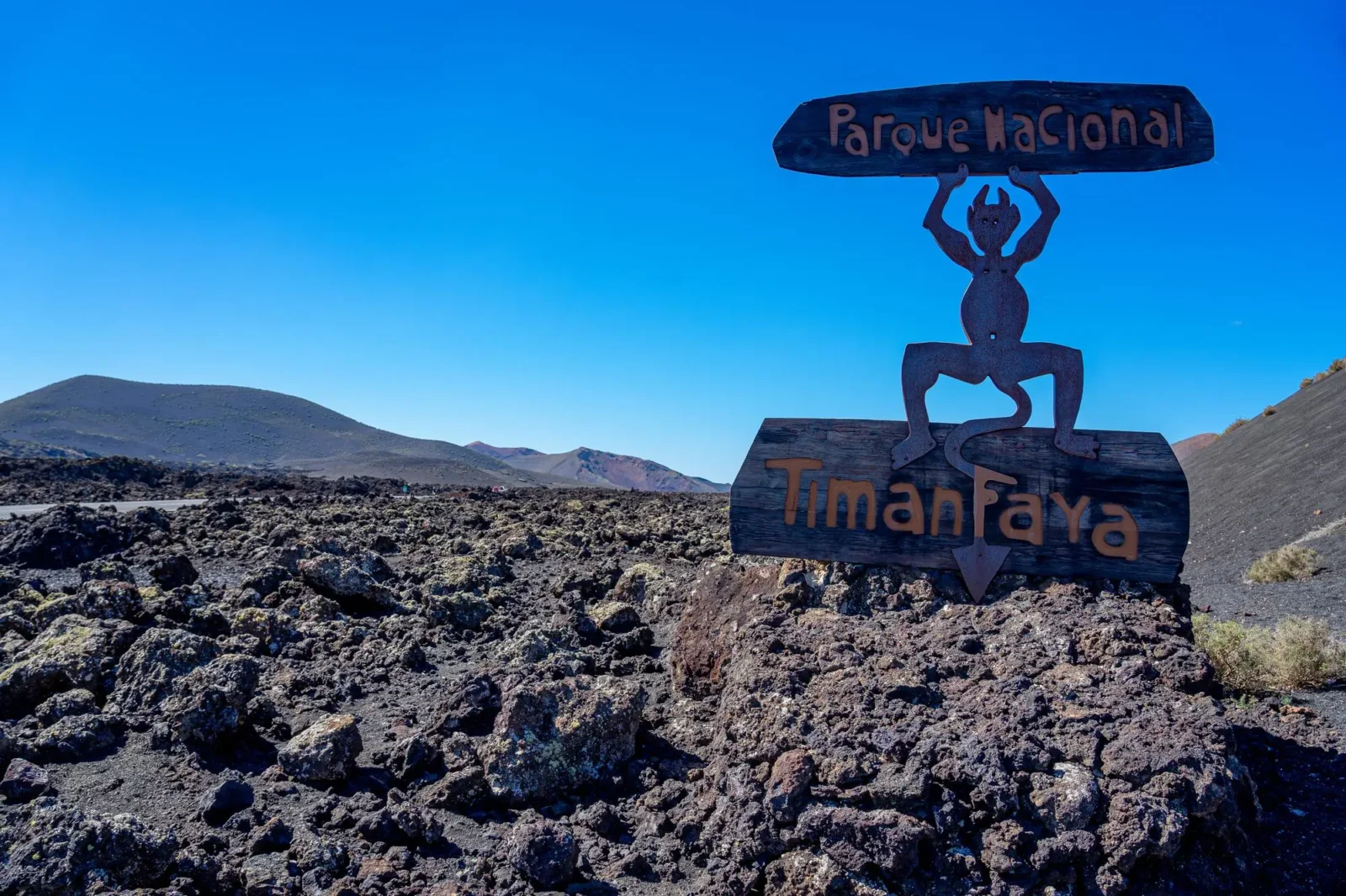Cedule Národního parku Timanfaya na Lanzarote obklopená černým lávovým polem a sopečnou krajinou pod jasně modrou oblohou.